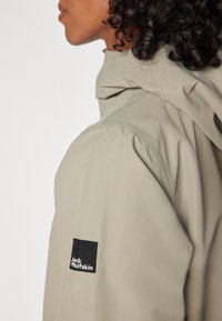 Béžová bunda s kapucňou, vyrobená z textúrovanej látky s naťahovacími rukávmi. Na rukáve je čierny štvorcový nášivok s logom "Jack Wolfskin".