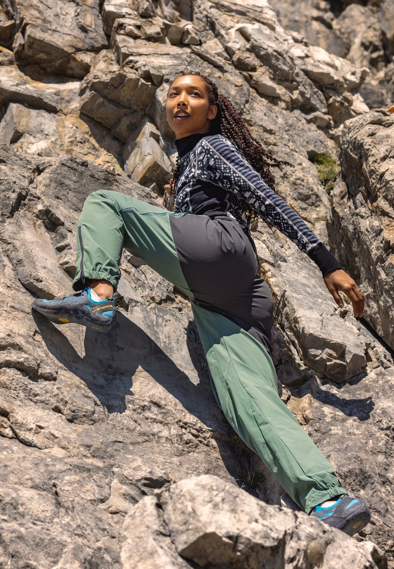 Grimpeur portant un haut à manches longues noir avec des manches à motifs, un pantalon vert et gris, et des chaussures d'escalade, grimpant sur une surface rocheuse.
