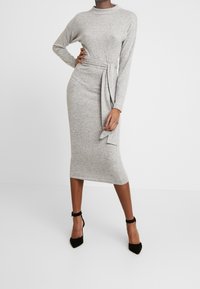 Femme portant une robe midi grise cintrée à manches longues avec une ceinture à la taille, associée à des escarpins noirs à brides à la cheville, debout devant un fond blanc.