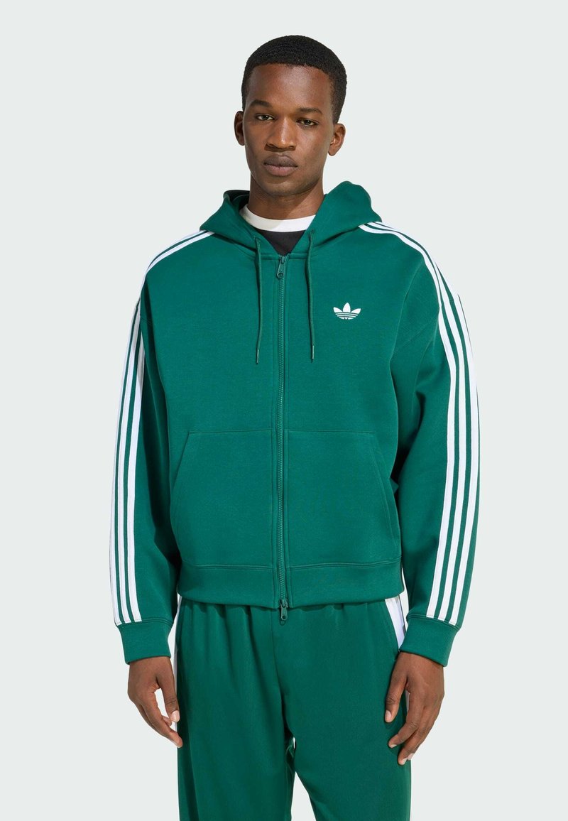 Felpa verde con zip realizzata in tessuto morbido, caratterizzata da strisce bianche Adidas sulle maniche e logo sul petto. Tasche a kangaroo sul davanti.
