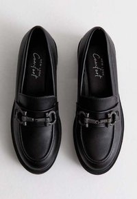 Mocasines de cuero negro estilo mocasín con una correa texturizada y hardware metálico decorativo. Presentan una suela plana y un diseño de punta redonda.