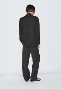 Grijze pinstripe blazer met een hoge kraag, gecombineerd met losse grijze broek, beide in een gestructureerde stof. Gestyled met zwarte slippers.