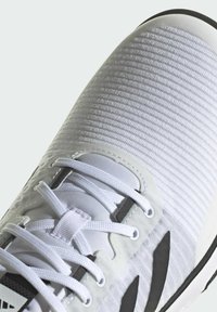 Weißer Sportschuh mit einer strukturierten, gerippten Oberseite, dünnen Schnürsenkeln und schwarzen Akzenten an den Seiten, ausgestattet mit einer gepolsterten Sohle und einem eleganten Design.