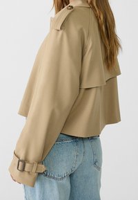 Krótka beżowa kurtka z szerokim kołnierzem, warstwowym krojem i regulowanymi paskami przy mankietach. Gładka tkanina o czystym wykończeniu, w połączeniu z niebieskimi dżinsami.