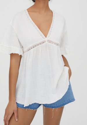 White, textured top with a deep V-neckline, lace detailing along the chest, and short, ruffled sleeves. Paired with denim shorts.