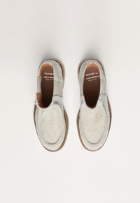 Chaussures slip-on en daim beige avec un bout ovale, semelles brunes contrastantes, et sans lacets ou fermetures visibles. L'étiquette intérieure indique "Woman by Fred Martin, fabriqué en Espagne."