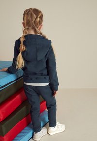 Set format din hanorac și pantaloni jogger bleumarin cu manșete reiate, asortat cu adidași albi. Părul copilului este aranjat în două codițe împletite.