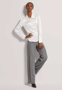 Vrouw die tegen de muur staat in een witte blouse met knopen, grijze wijde broek en zwarte hakken, met één hand op de heup poserend.