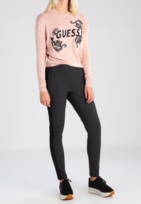 Chemise rose clair à manches longues avec "GUESS" en sequins noirs ; associée à un pantalon noir ajusté et des baskets noires avec une semelle blanche.