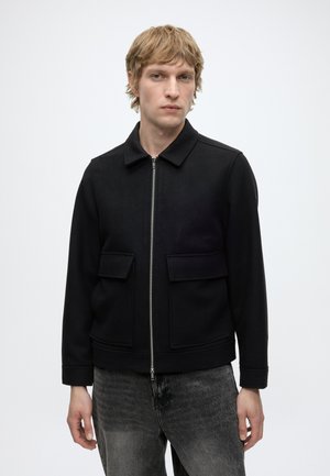 Junger Mann mit blondem Haar, der eine schwarze Jacke mit Reißverschluss und Frontklapptaschen sowie ausgewaschene schwarze Jeans trägt, steht vor einem schlichten Hintergrund.