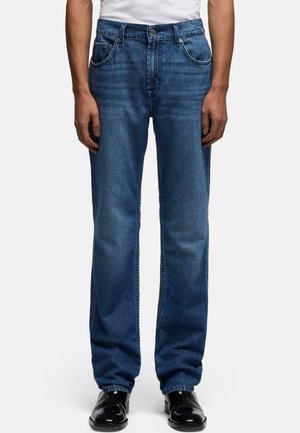 Homme portant un jean bleu droit avec poches avant et des chaussures habillées noires, debout devant un fond clair uni.