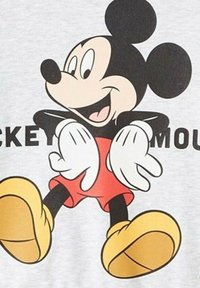 T-shirt gris avec un graphique de Mickey Mouse dans des couleurs noire, rouge et jaune. La souris a de grandes oreilles, des gants blancs et de grandes chaussures.