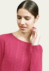 Pull en tricot câblé fuchsia avec un col rond et des poignets côtelés, présentant un motif texturé sur le devant.