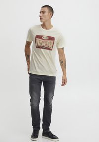 Tricou crem cu un grafic vintage roșu, pe care scrie "BLEND VINTAGE". Purtați cu blugi gri închis și adidași negri. Fundal simplu.