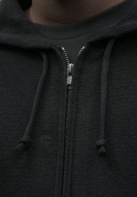 Czarna bluza z kapturem na zamek, wykonana z teksturowanej tkaniny, z metalowym zamkiem błyskawicznym i detalami ze sznurkiem. Zbliżenie na linię szyi i obszar zamka.