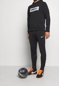 Černá mikina Nike s bílým logem, černé přiléhavé kalhoty a černé fotbalové boty s oranžovými akcenty. Vedle leží černý fotbal.