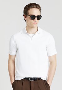 Polo shirt bianco a maniche corte con un motivo texturizzato, dotato di colletto. Il modello indossa occhiali da sole neri e pantaloni marroni con una cintura nera.