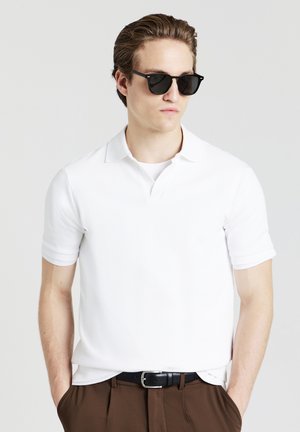 Polo shirt bianco a maniche corte con un motivo texturizzato, dotato di colletto. Il modello indossa occhiali da sole neri e pantaloni marroni con una cintura nera.