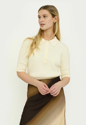 Pull en maille polo à manches courtes crème avec un motif texturé et un col à boutons, associé à une jupe en satin dégradé marron.