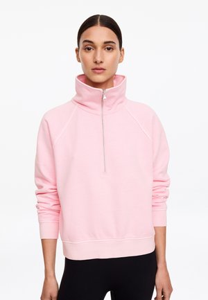 Femme portant un sweat à capuche zippé rose clair avec un col haut et des manches longues, debout devant un fond blanc.