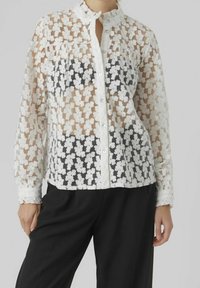 Blouse blanche transparente à motif floral, manches longues, boutonnée sur le devant, avec un col haut, portée sur un haut sombre. Tissu texturé.