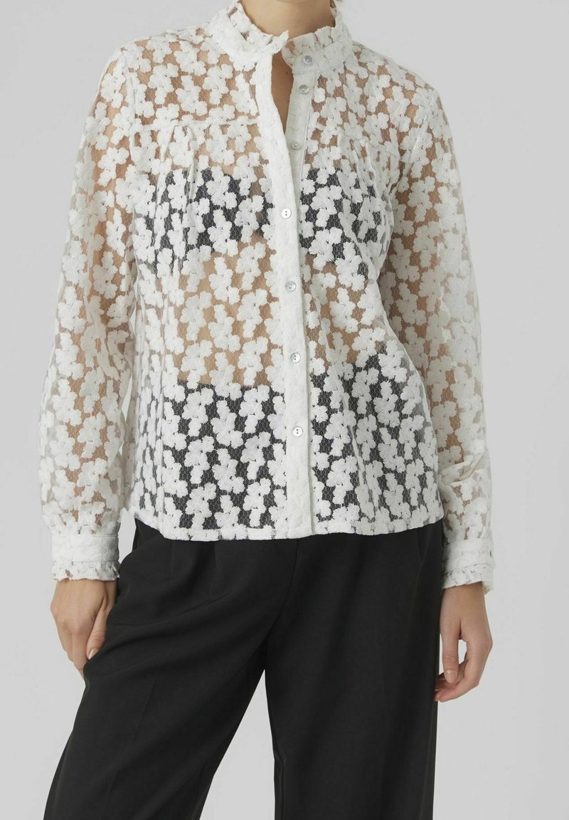Blouse blanche transparente à motif floral, manches longues, boutonnée sur le devant, avec un col haut, portée sur un haut sombre. Tissu texturé.
