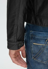 Schwarze Lederjacke mit Druckknopfdetail an den Bündchen, getragen über blauen Jeans mit kontrastierenden Nähten und einem Design für die Gesäßtasche.