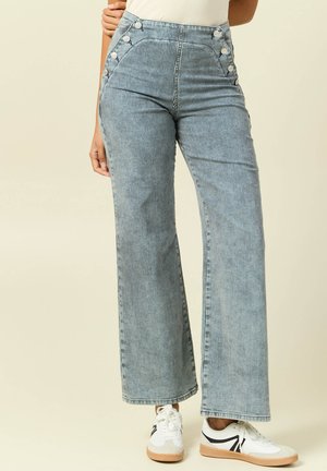 Vaqueros de talle alto y pierna ancha con finas rayas verticales y detalles de botones laterales, combinados con zapatillas blancas.