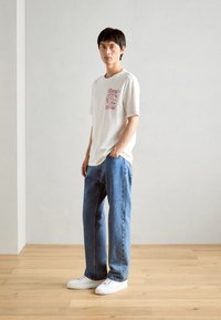 Creme graphic t-shirt med rød print, parret med løst siddende lyseblå denimjeans og hvide sneakers, sat mod en neutral baggrund.