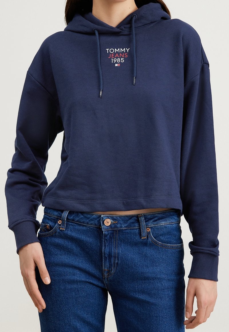 Křácená námořnicky modrá mikina s kapucí na šňůrku, zdobená malým červeno-bílým logem. Kombinováno s modrými džínami z denimu.