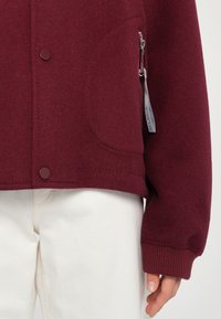 Bordeaux fleece jack met een ronde zak, ritsdetail en ribbelmanchetten. Gecombineerd met lichtgekleurde broeken.