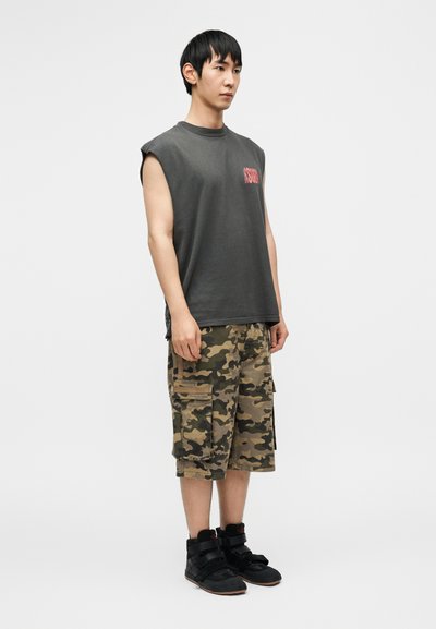 Αμάνικη σκουρόγρια μπλούζα με κόκκινα γράμματα, σε συνδυασμό με καμουφλάζ βερμούδα cargo με πολλές τσέπες και άνετη εφαρμογή, και μαύρα ψηλά αθλητικά παπούτσια.