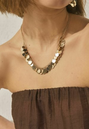 Woman wearing a gold chain necklace with round reflective discs and matching earrings, dressed in a strapless brown top.