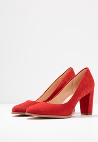 Escarpins à talons hauts en daim rouge avec un bout arrondi et un talon épais, dotés d'une doublure intérieure lisse et d'une surface extérieure texturée.