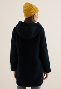 Tummansininen fleece-takki hupulla, jossa on teksturoitu kangas, ylisuuri mitoitus ja ribbikuvioinen sinapinkeltainen neulottu pipo. Mukava muotoilu lämpimyyteen.