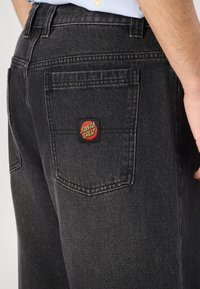 Črne jeans hlače iz denima z teksturiranim zaključkom, ki imajo rdečo in rumeno nalepko Santa Cruz na zadnjem žepu ter tradicionalno zasnovo žepov.