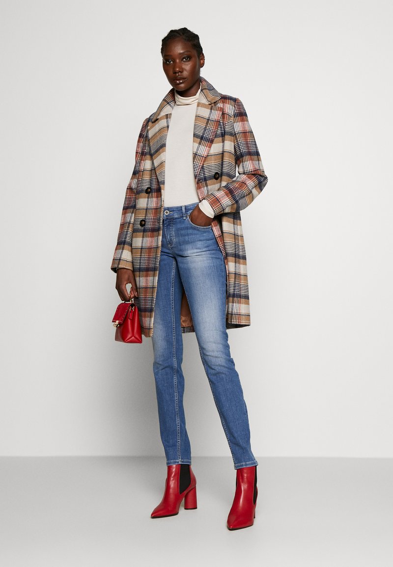 Kobieta w beżowym i granatowym w kratkę płaszczu, białym golfie, niebieskich dżinsach, czerwonych botkach na kostkę, trzymająca małą czerwoną torebkę, stoi pod białą ścianą.