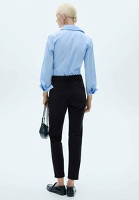 Svetlo modra prugasta srajca, črne hlače z visokim pasom in črne loafer čevlje. Model drži majhno črno torbico, gledano s hrbta na svetli podlagi.