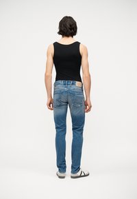 Άνδρας με μαλλιά μεσαίου μήκους, φοράει στενό μαύρο αμάνικο τοπ, ξεθωριασμένο μπλε τζιν και λευκά αθλητικά, γυρισμένος πλάτη σε απλό λευκό φόντο.