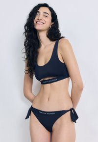 Σετ μπικίνι σε ναυτικό μπλε χρώμα με crop τοπ που έχει ανοίγματα και λωρίδα με λογότυπο, συνδυασμένο με σλιπ χαμηλής μέσης με κορδόνια. Λείο ύφασμα.