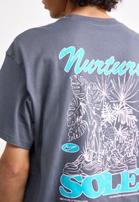 Tumši pelēks kokvilnas T-krekls ar tirkīza grafiku, kas lasāma "Nurture", un detalizētām baltām ilustrācijām par augiem un kedām mugurpuses. 