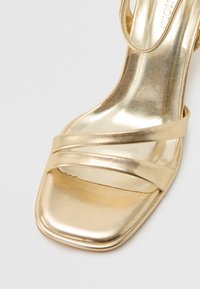 Χρυσό μεταλλικό πέδιλο με ανοιχτά δάχτυλα και τετράγωνη μύτη, δύο λεπτά σταυρωτά λουριά πάνω στα δάχτυλα και ένα μόνο λουράκι στον αστράγαλο.