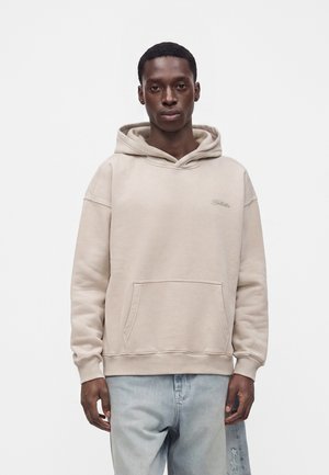 Junger Mann, der einen beigen Hoodie und hellblaue Jeans trägt, steht frontal vor einem schlichten weißen Hintergrund.