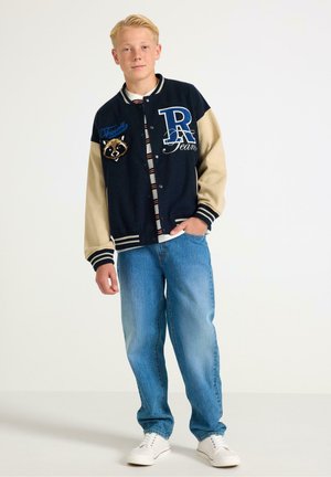 Navy and beige varsity jacket with embroidered raccoon, striped button-up shirt underneath, paired with light blue jeans and white sneakers.