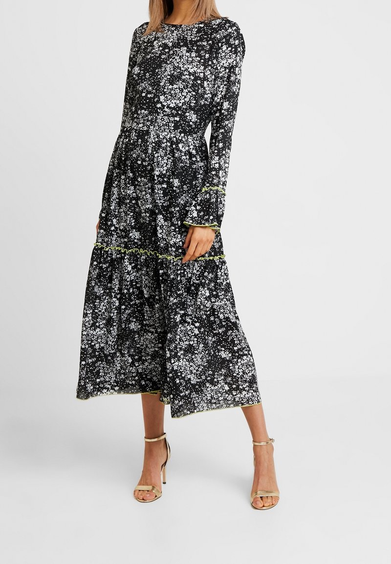Femme portant une robe midi noire à imprimé floral blanc, manches longues, bordure volantée vert lime et sandales à talons beiges.
