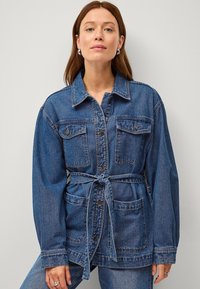 Giacca di jeans blu medio, con maniche lunghe, tasche con bottone e cintura da annodare in vita con una finitura testurizzata.