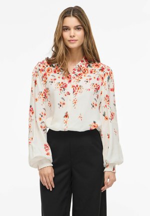 Nő fehér, hosszú ujjú blúzt visel piros és narancssárga virágmintával, magasderekú fekete nadrágba betűrve, fehér háttér előtt.