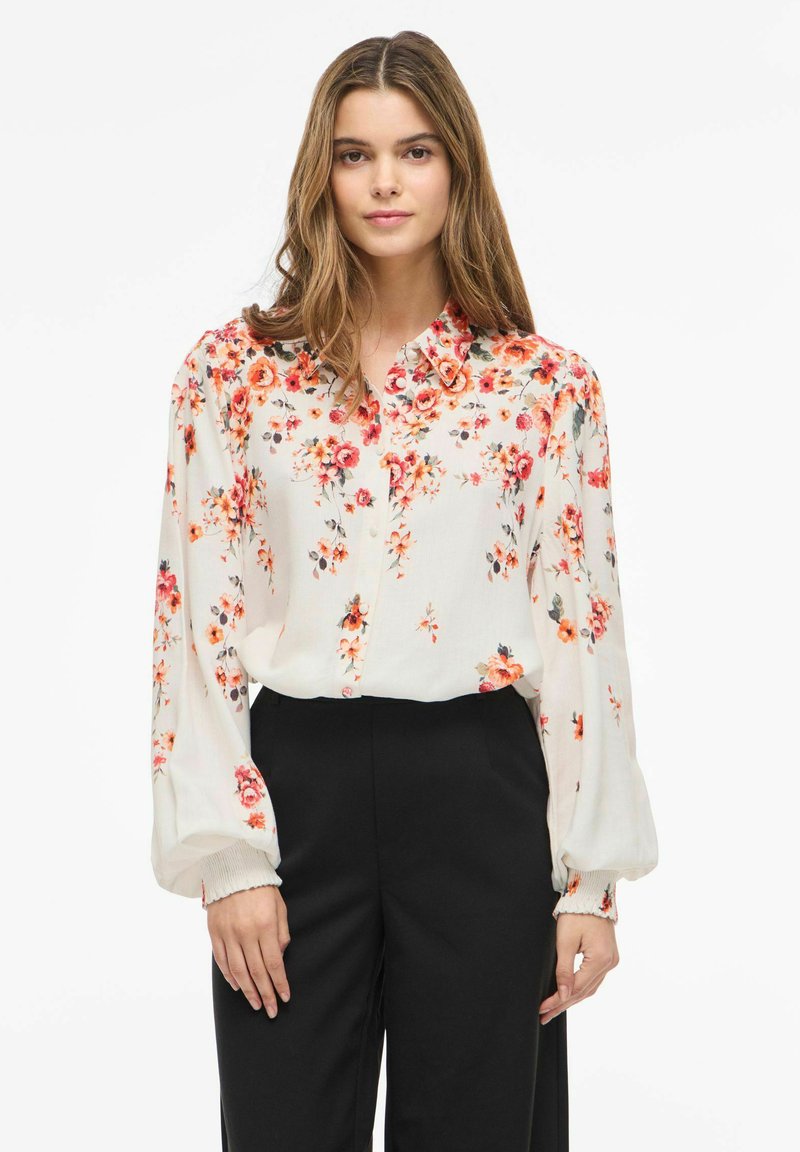 Femme portant un chemisier blanc à manches longues avec un imprimé floral rouge et orange, rentré dans un pantalon noir taille haute sur fond blanc.