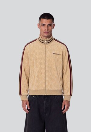 Uomo che indossa una giacca beige con zip con texture a motivo e strisce bordeaux sulle maniche, abbinata a pantaloni neri larghi, in piedi contro uno sfondo grigio.