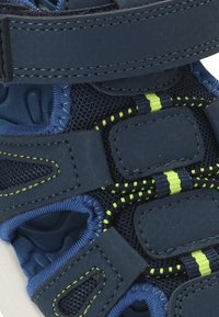 Primer plano de una sandalia deportiva con correas azul oscuro, velcro ajustable y costuras amarillas brillantes sobre un fondo de malla texturizada.